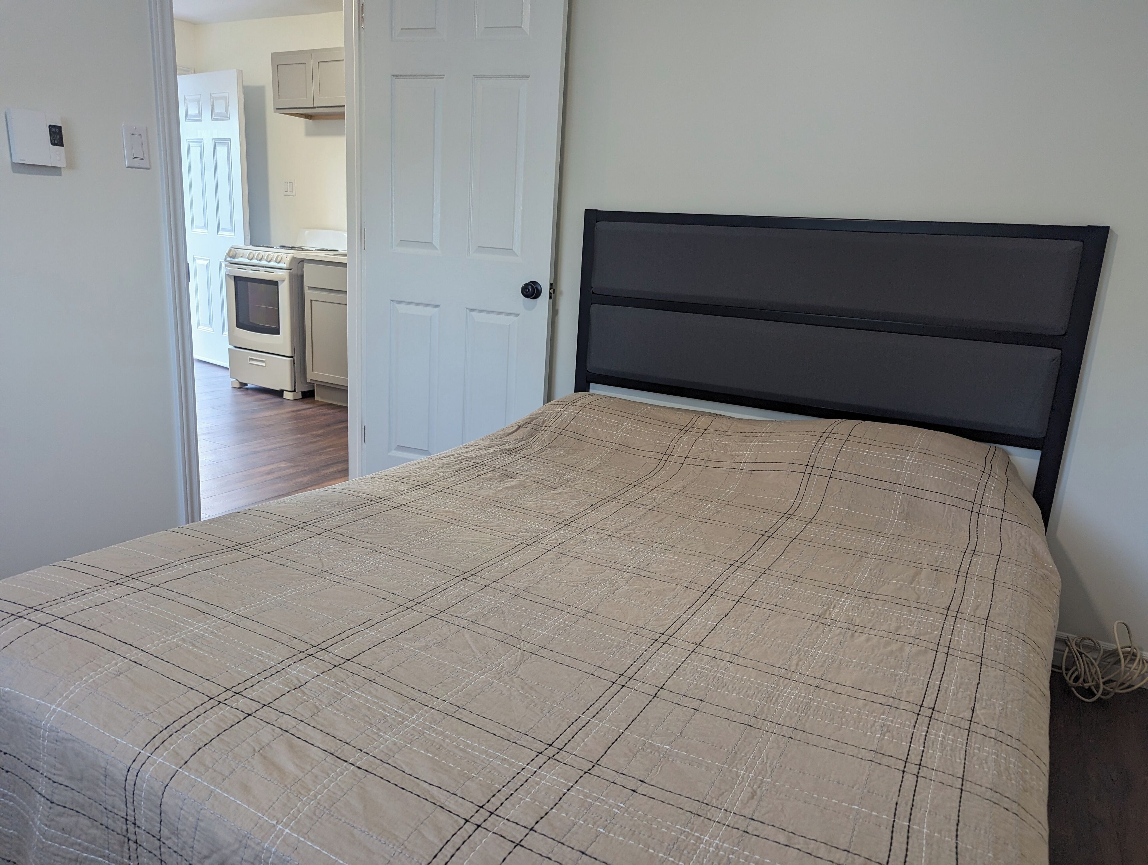 A bedroom with a bed, a door, and a view into a kitchen area.