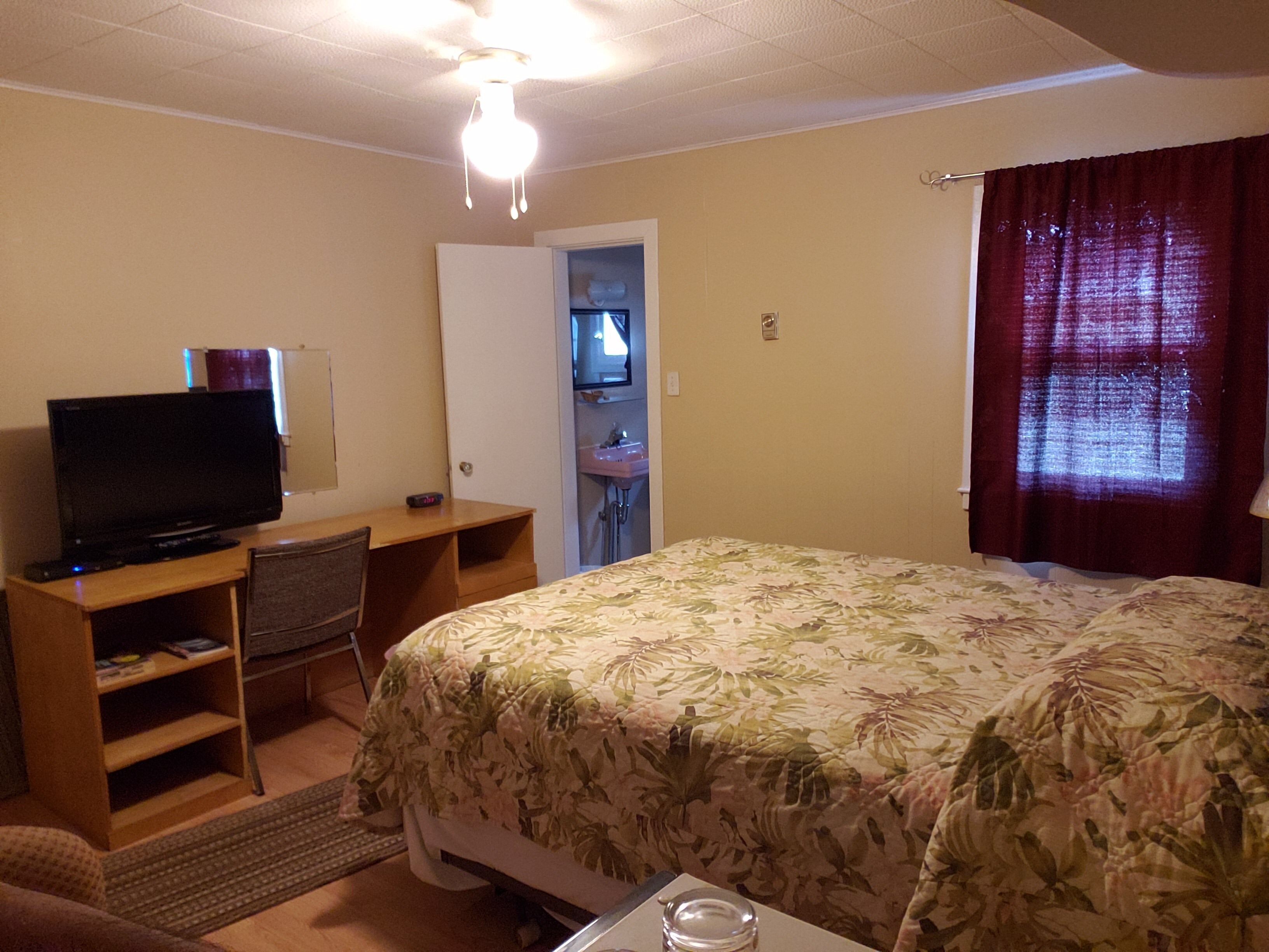 A hotel room with a bed, a desk with a television, a chair, a small table, and a bathroom visible through an open door.