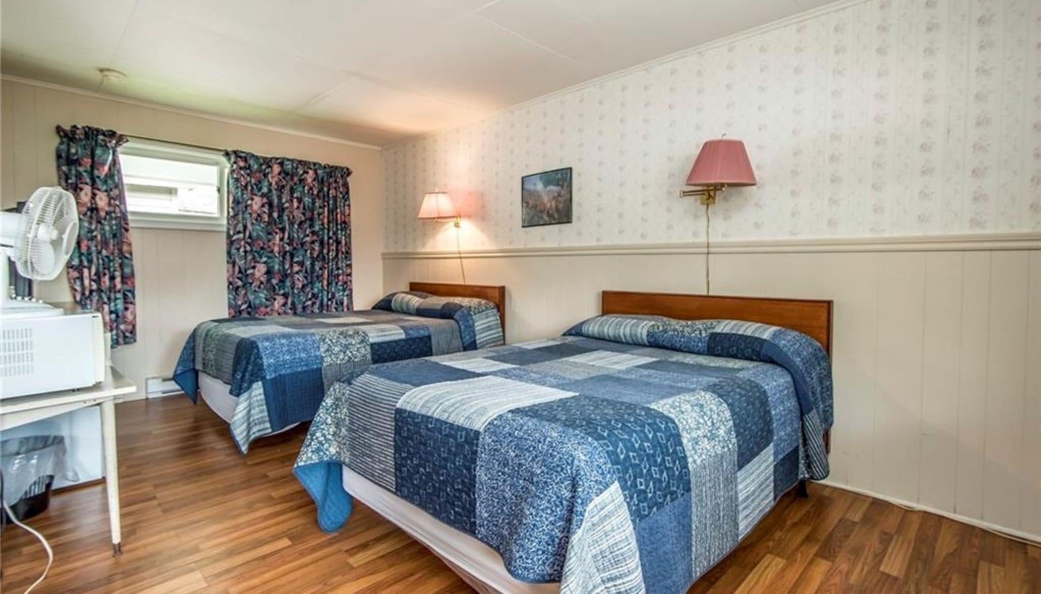 A room with two beds, a desk, a lamp, and a fan.
