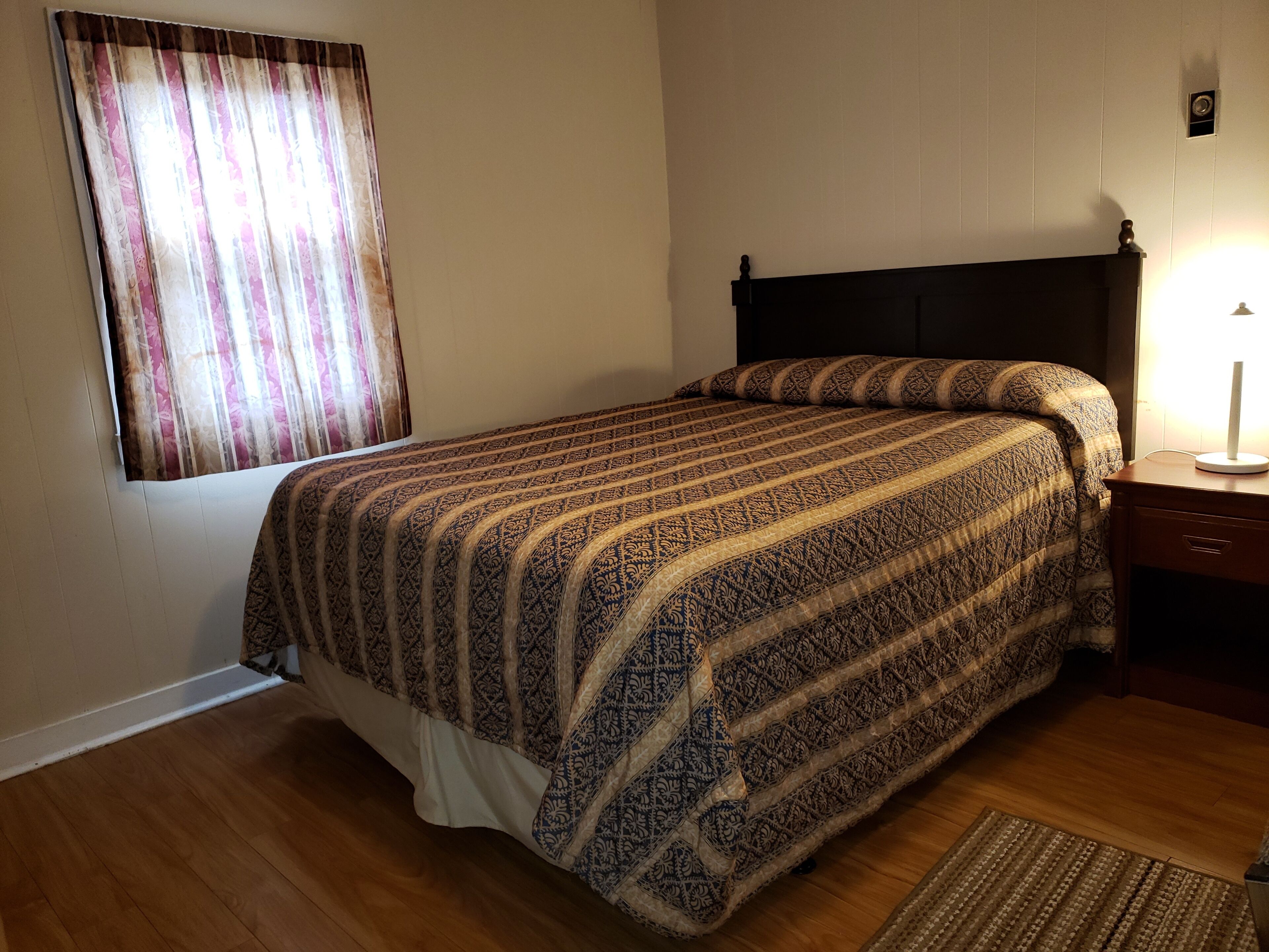 Une chambre à coucher avec un lit, un plancher en bois, une table de chevet, une lampe et une fenêtre avec des rideaux.
