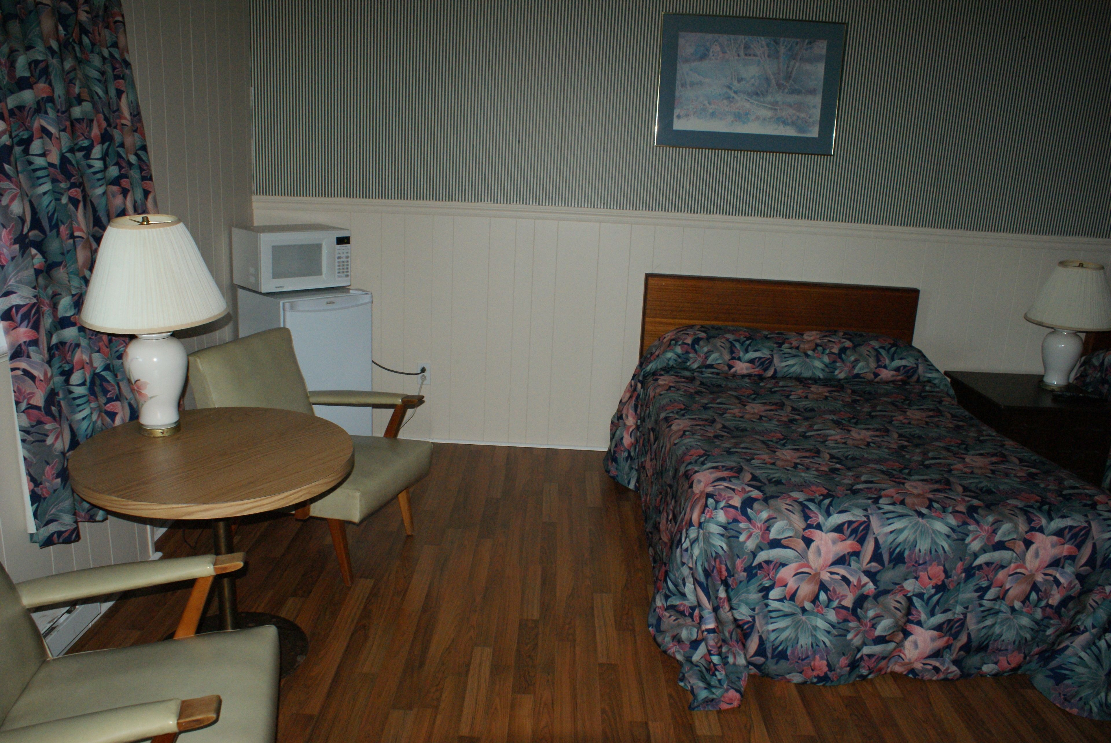 A hotel room with a bed, a small table with a lamp, a chair, a microwave, and a refrigerator.