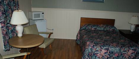 A hotel room with a bed, a small table with a lamp, a chair, a microwave, and a refrigerator.