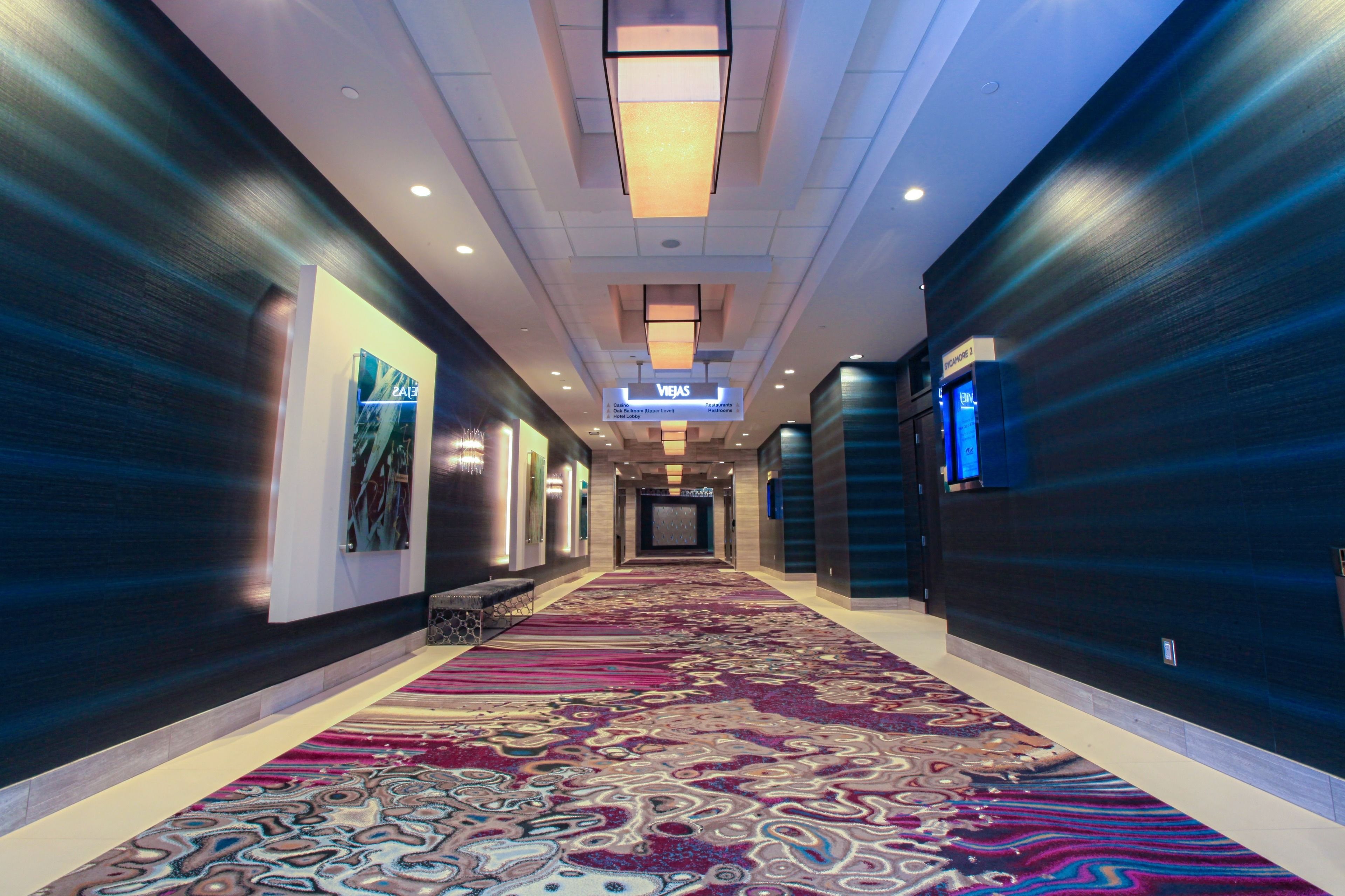 Pasillo de hotel con alfombra estampada, paredes oscuras y un letrero que indica la dirección a los ascensores.