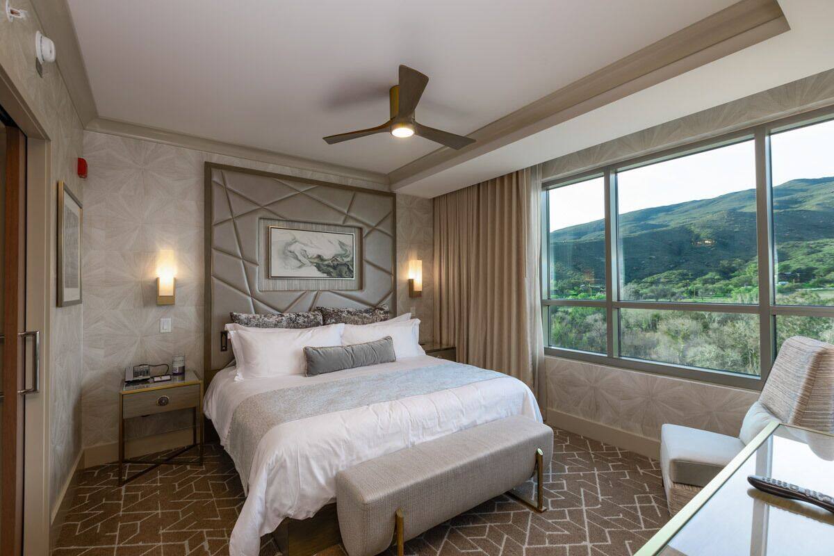 Une chambre d’hôtel avec un grand lit, un bureau avec une chaise, un ventilateur de plafond et une grande fenêtre donnant sur des collines verdoyantes.