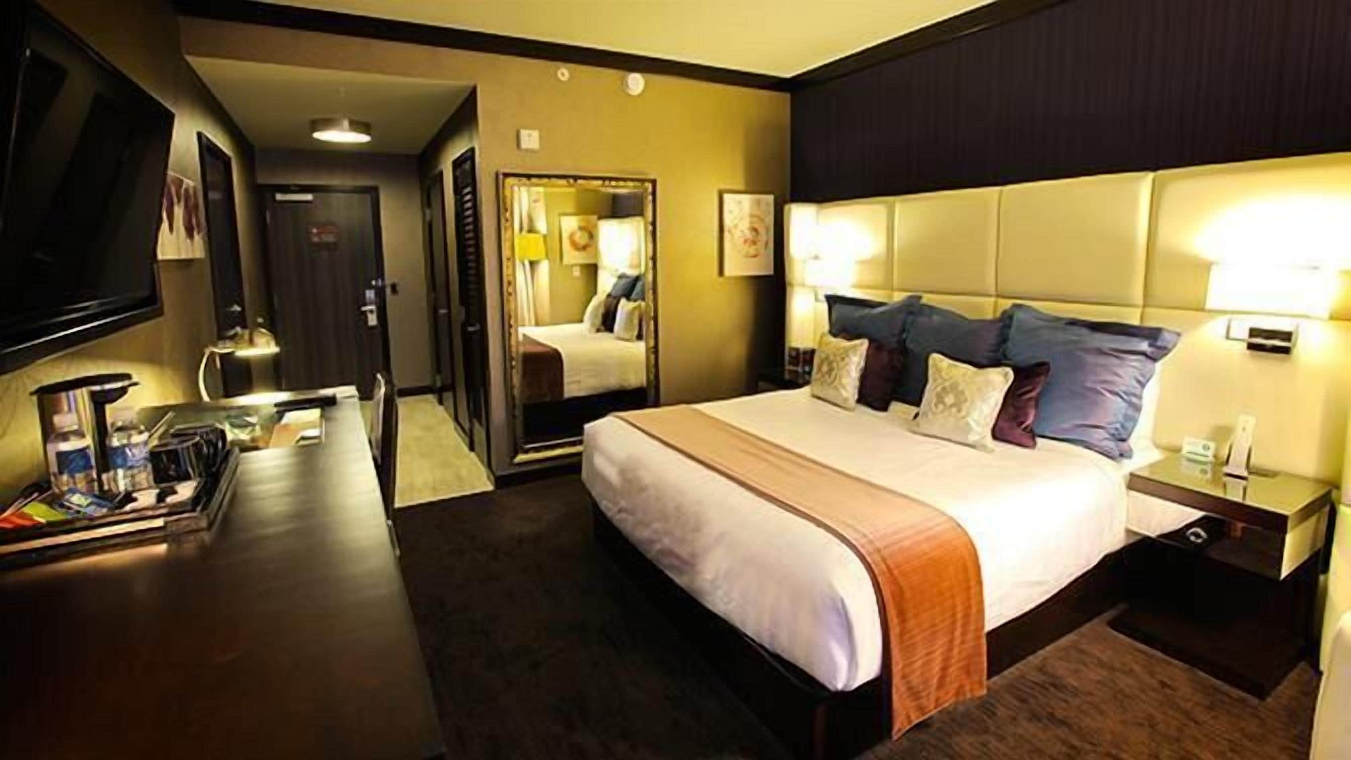 Habitación de hotel con una cama grande, mesitas de noche, un escritorio con silla, un televisor de pantalla plana y un espejo.