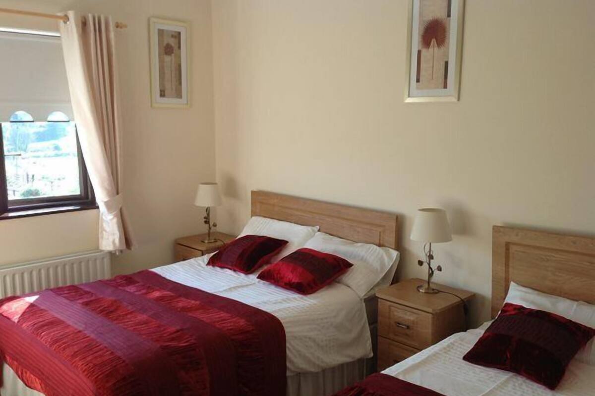 A hotel room with two beds, each with a wooden headboard, white bedding, and red pillows. There are bedside tables with lamps, a window with curtains, and a radiator.