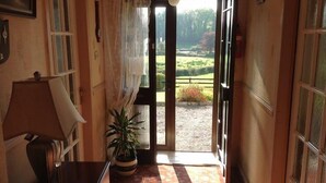 A hallway with a wooden floor, a potted plant, a lamp, and a door leading to an outdoor area with a view of greenery.