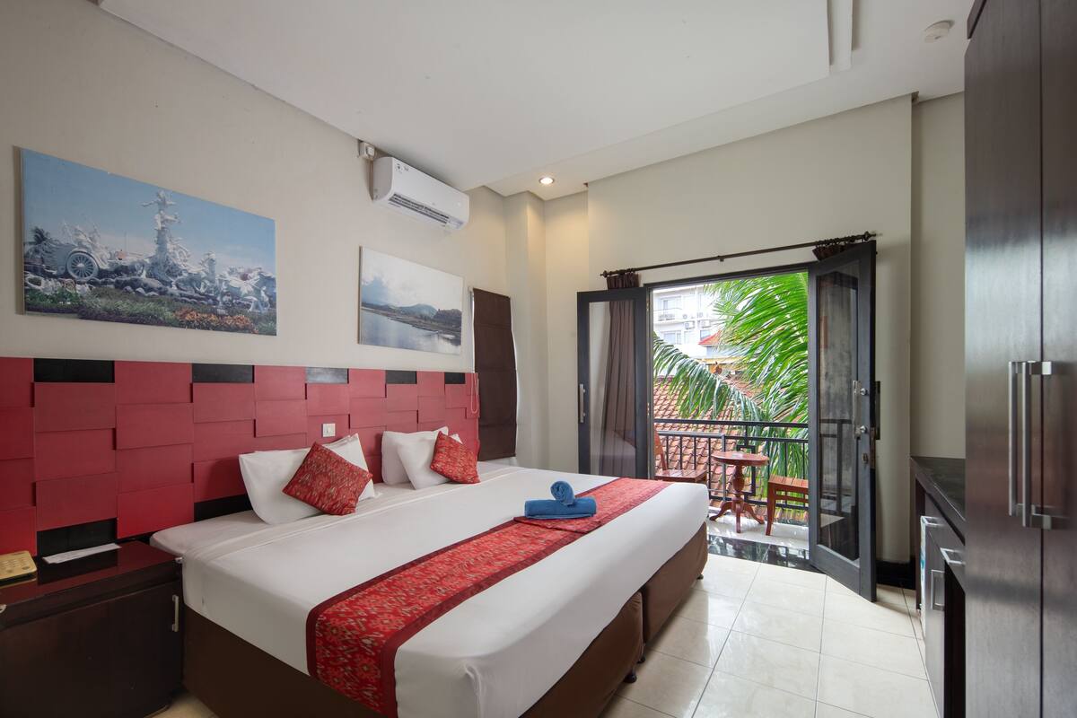 A hotel room with a large bed, a red headboard, a balcony with a view of greenery, and a kitchen area.