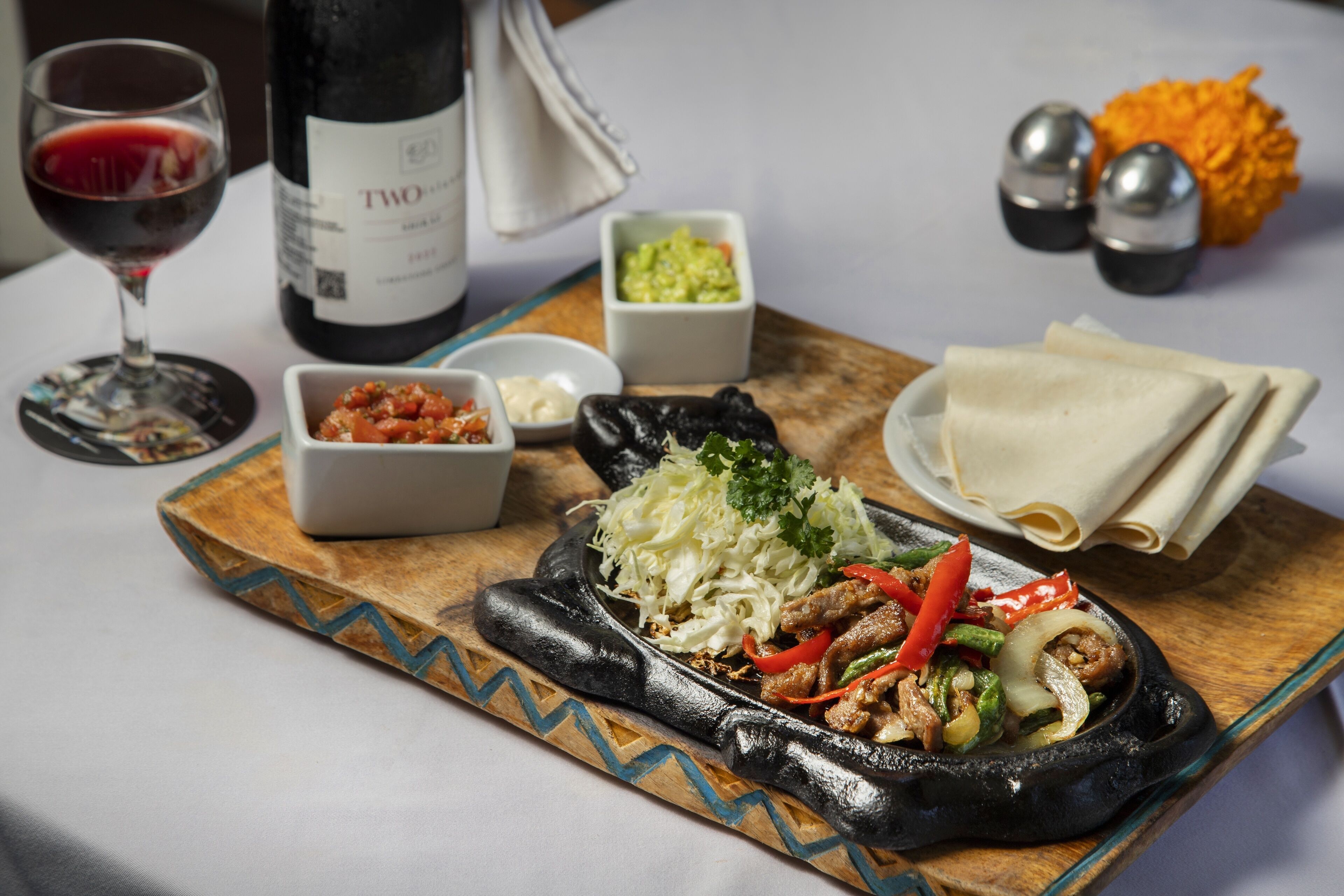 A meal with a black skillet, vegetables, and meat, accompanied by a bottle of wine, bowls of condiments, and a napkin on a wooden board.