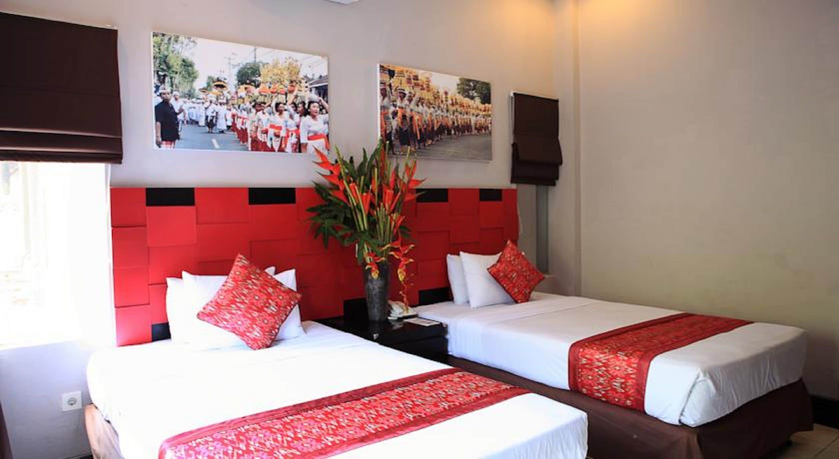 A hotel room with two beds, red headboard panels, white bedding, and a vase with red flowers.