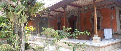Ein traditionelles Gebäude mit einem Ziegeldach und einer Veranda. Im Vordergrund befinden sich Kübelpflanzen und ein Baum.