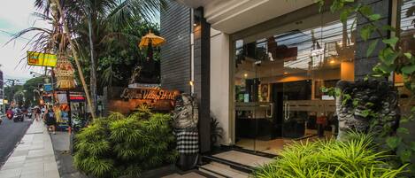 A hotel entrance with a sign reading "Legia Village Hotel," a tiled sidewalk, and lush greenery.