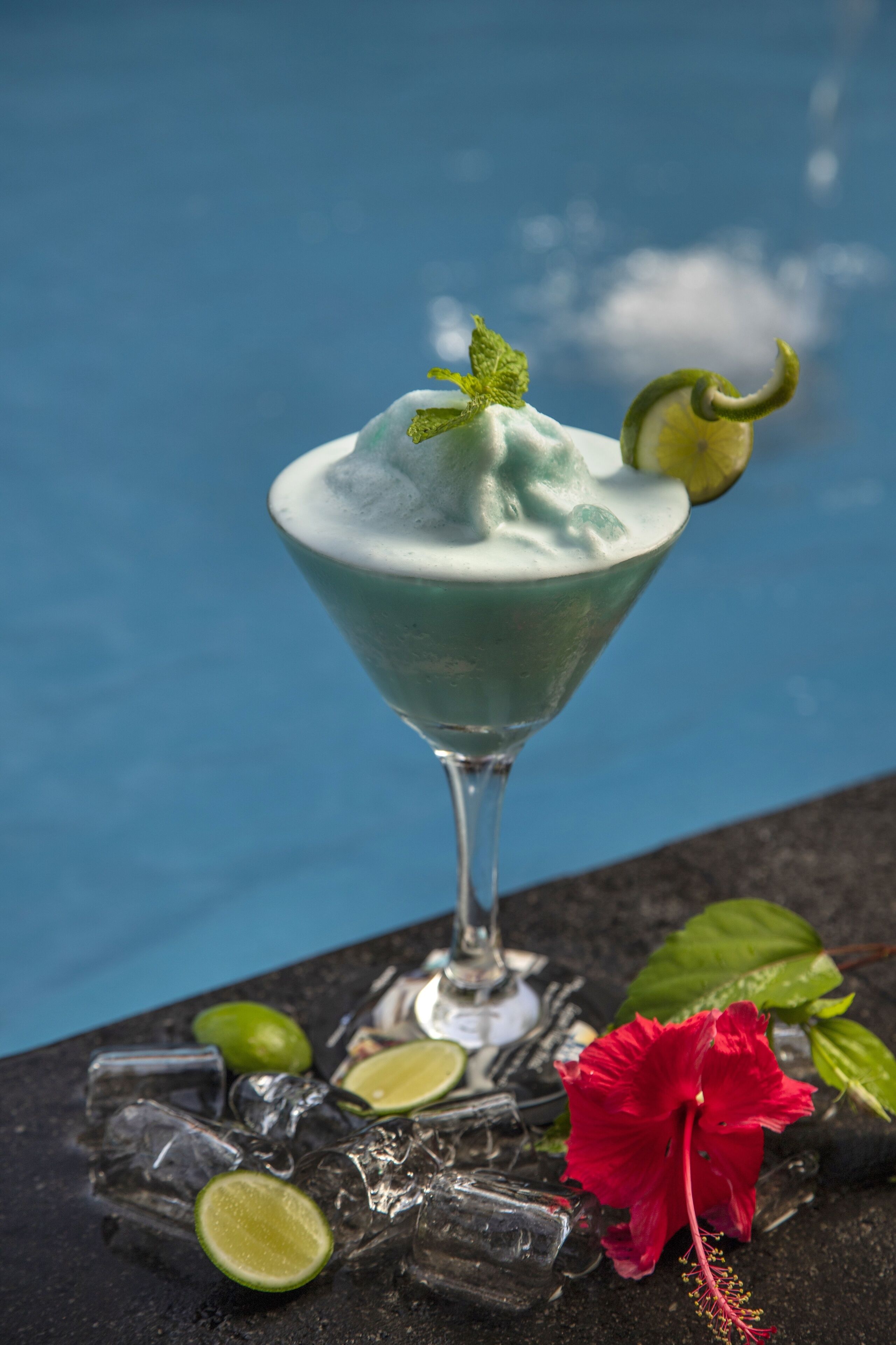 Un cóctel en una copa de martini, adornado con una rodaja de lima, sobre una mesa con cubitos de hielo y una flor de hibisco rojo.