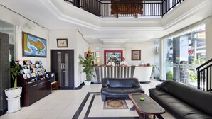 A hotel lobby with a reception desk, black leather sofas, a coffee table, a potted plant, and a large window offering a view of the outside. - Legian Village Hotel (Legian)