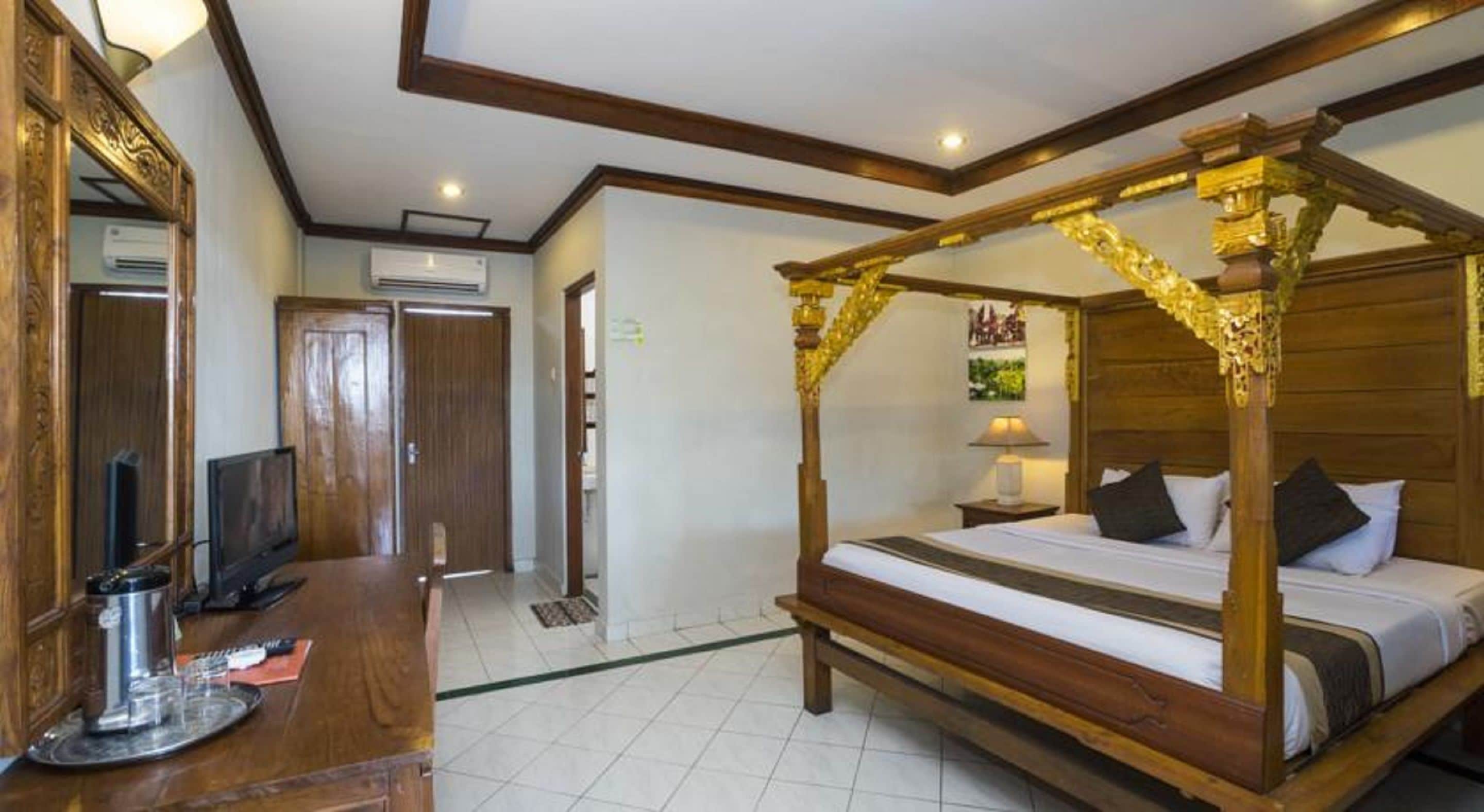 A four-poster bed with a wooden frame and gold accents, a television on a wooden desk, and a door leading to another room.