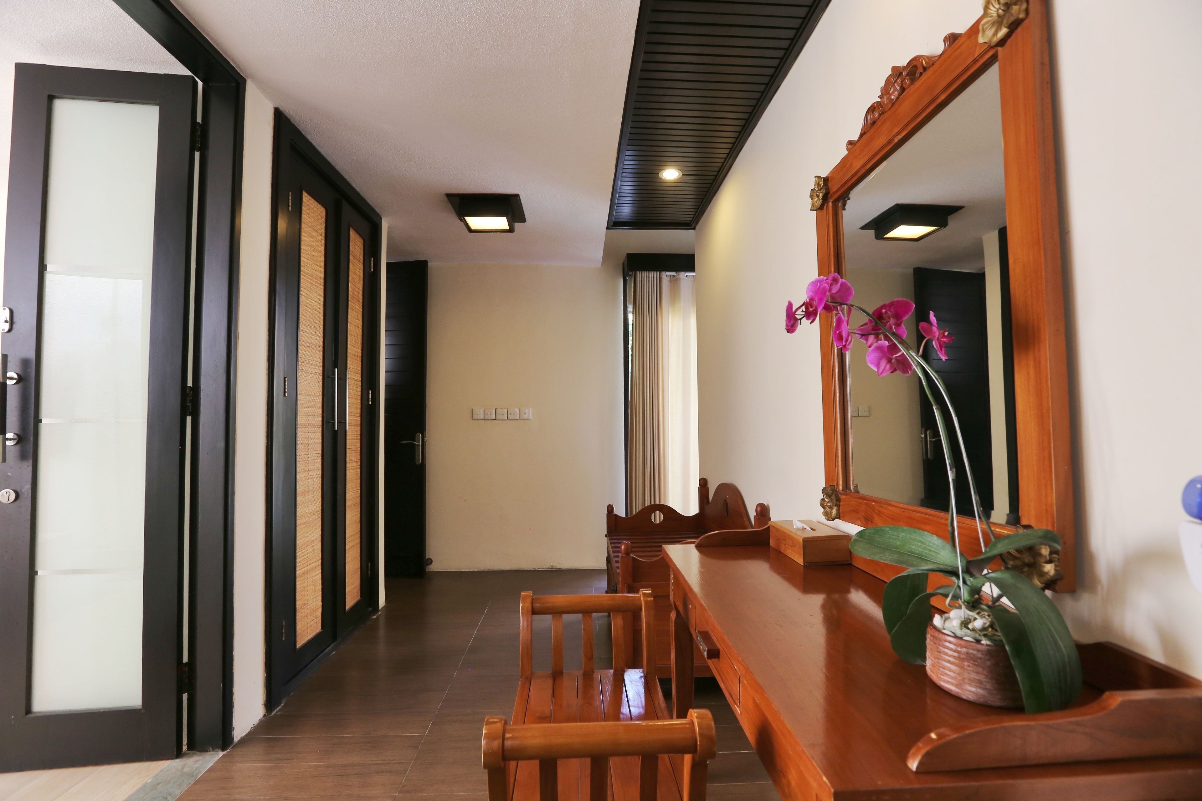 Un couloir avec un banc en bois, une table à manger et un grand miroir qui reflète une orchidée en pot.