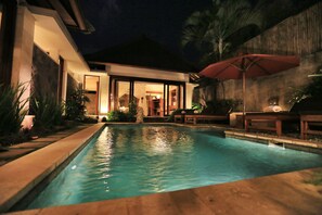 Une piscine avec une terrasse en bois, un parasol rouge et une maison moderne dotée de portes vitrées.