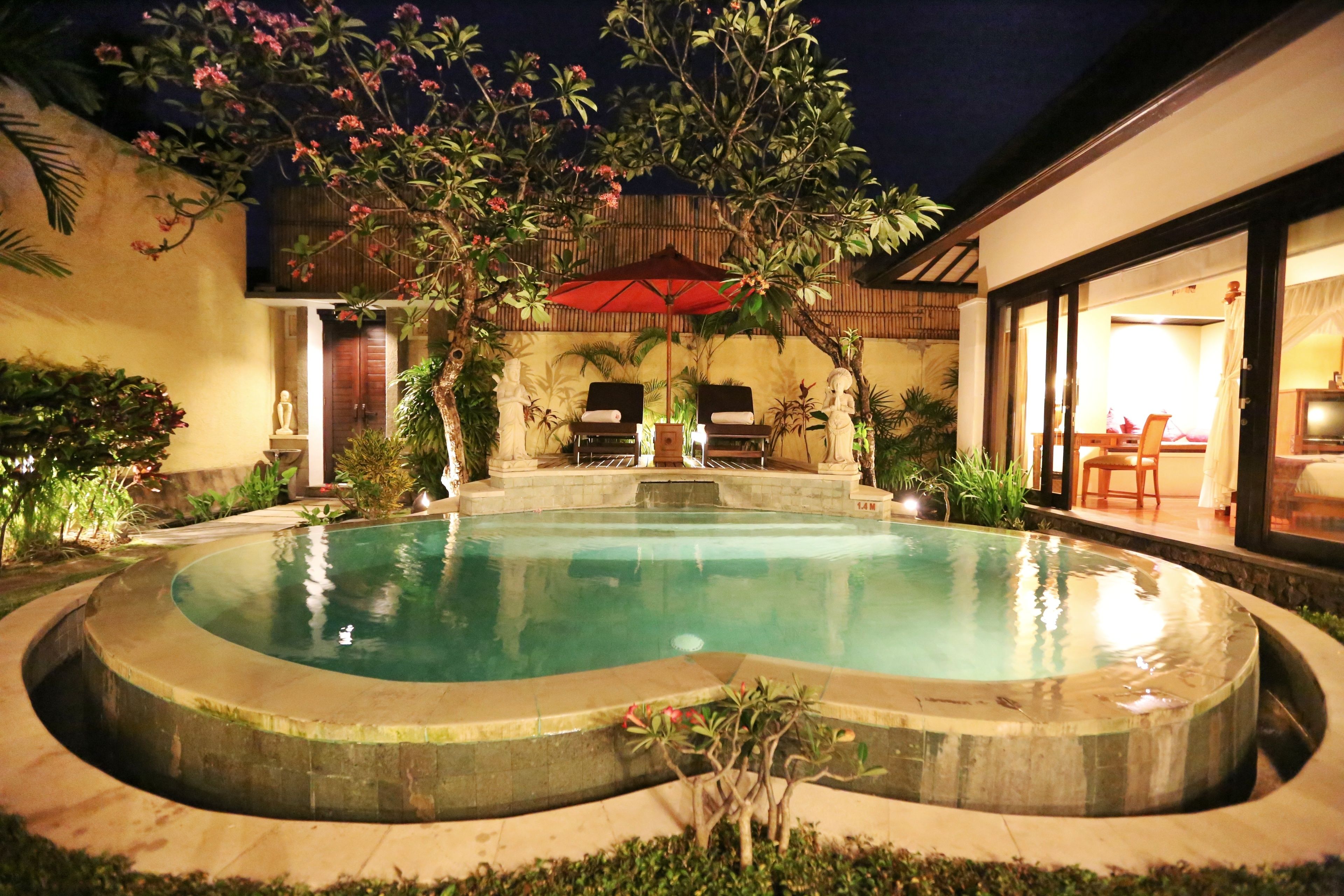 A circular pool with a stone edge, surrounded by lush greenery and a wooden deck. There are two lounge chairs under a red umbrella.