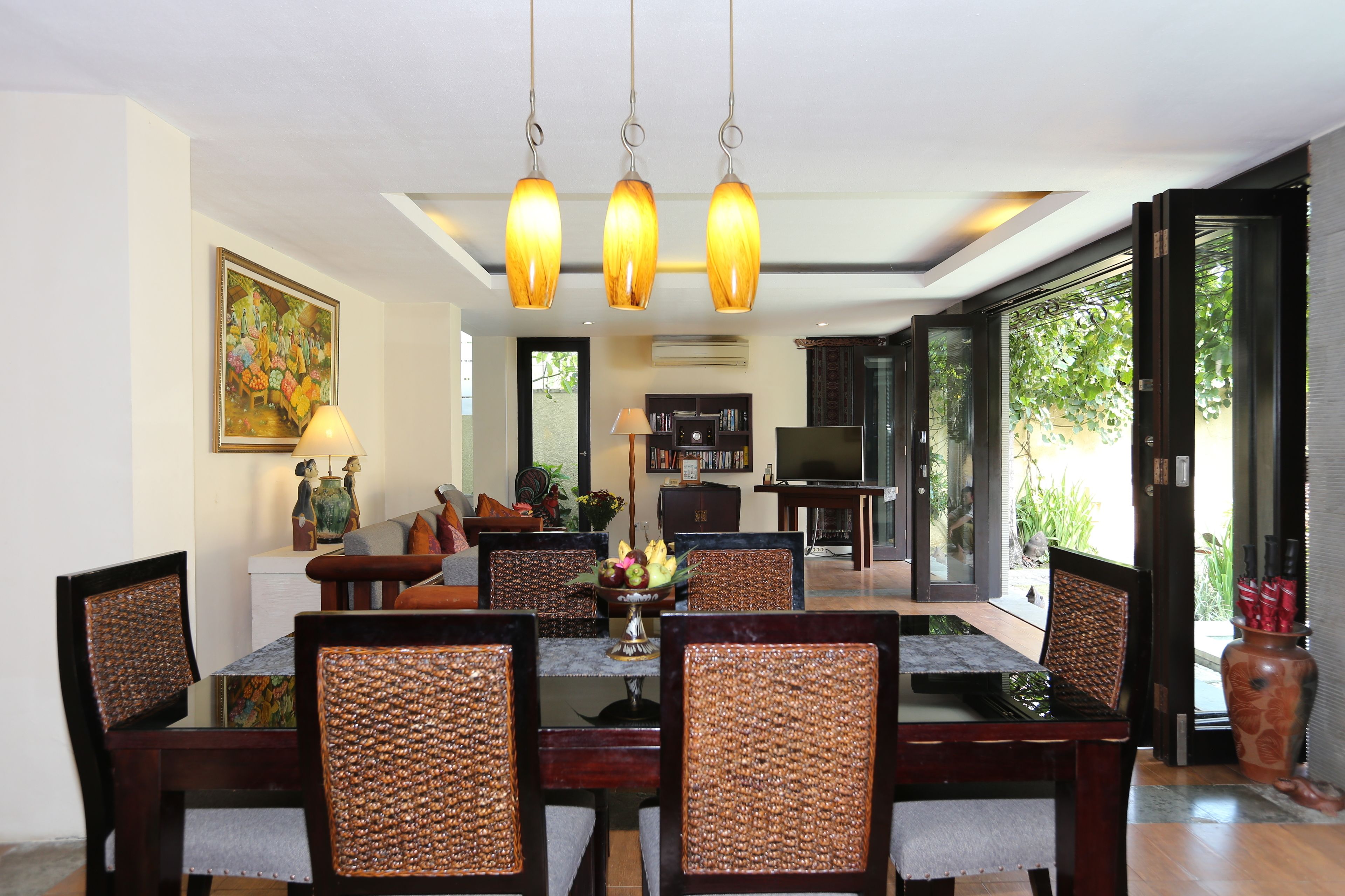 A dining area with a table set for six, surrounded by wicker chairs, a chandelier, and a bookshelf in the background.