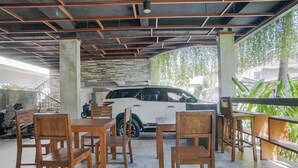 An outdoor seating area with wooden tables and chairs, a white car parked nearby, and a view of greenery. - Akarsa Kokoro Sanur Bali (Denpasar)