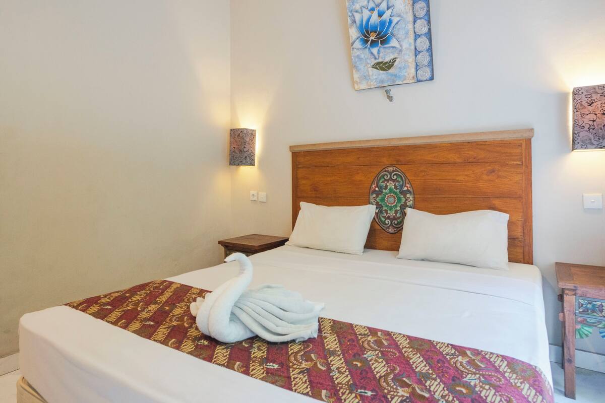 A neatly made bed with a patterned bedspread and a swan-shaped towel. There are two wall-mounted lamps and a decorative headboard.