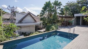 A swimming pool with a tiled deck, surrounded by greenery and a thatched-roof building. - Akarsa Kokoro Sanur Bali (Denpasar)