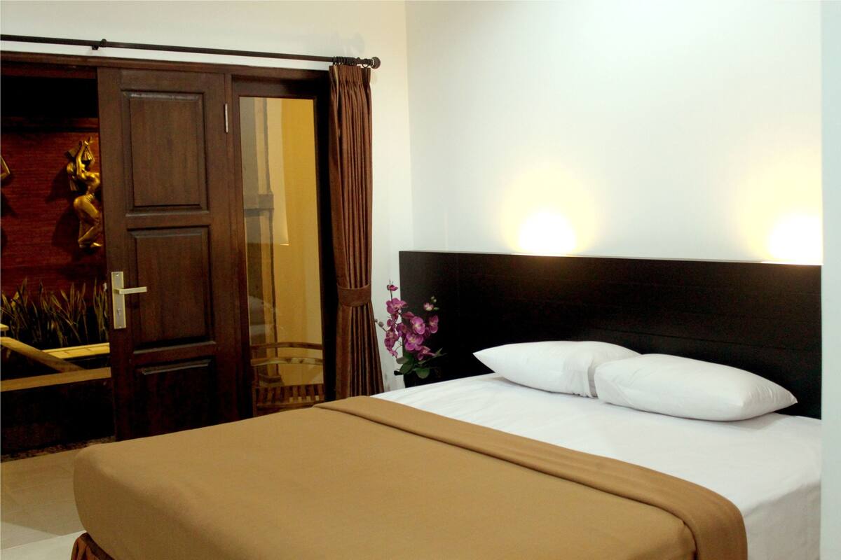 Une chambre d’hôtel avec un lit, une porte en bois et une table de chevet sur laquelle se trouve un vase de fleurs.