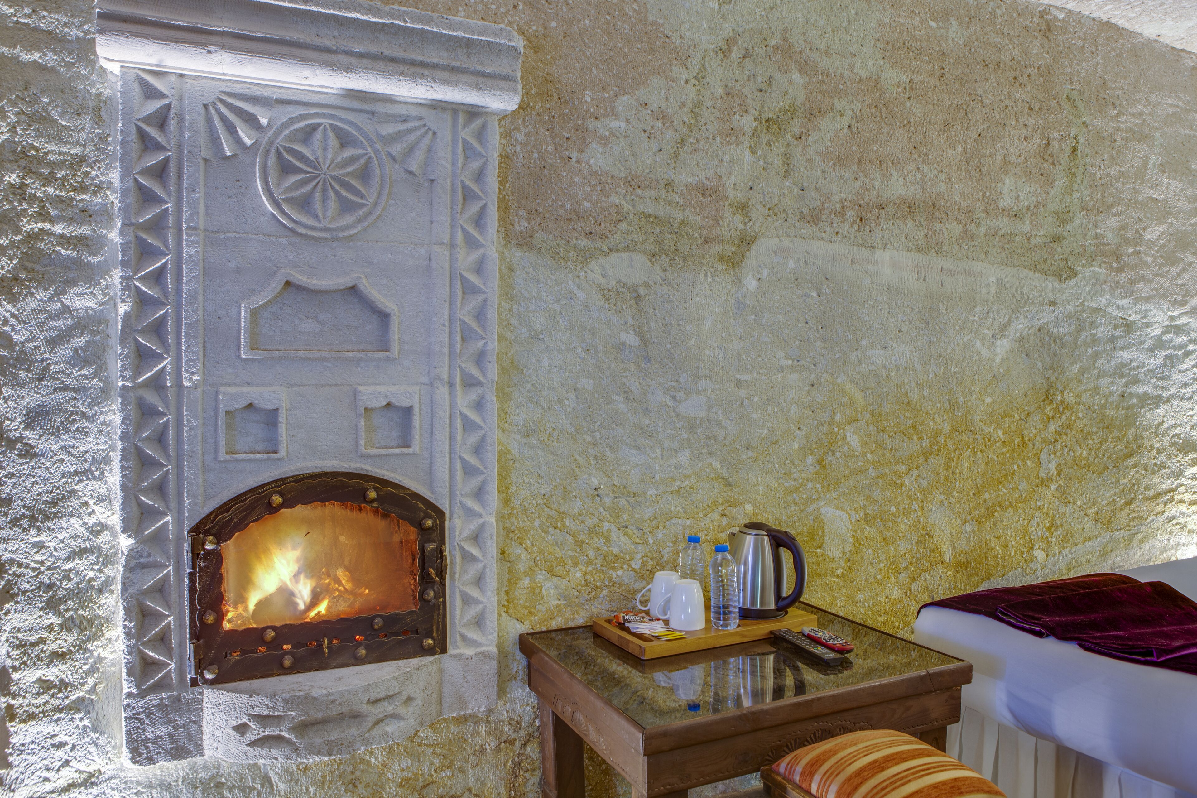 A rustic room with a stone fireplace, a wooden table with a tray of items, and a bed with a purple blanket.