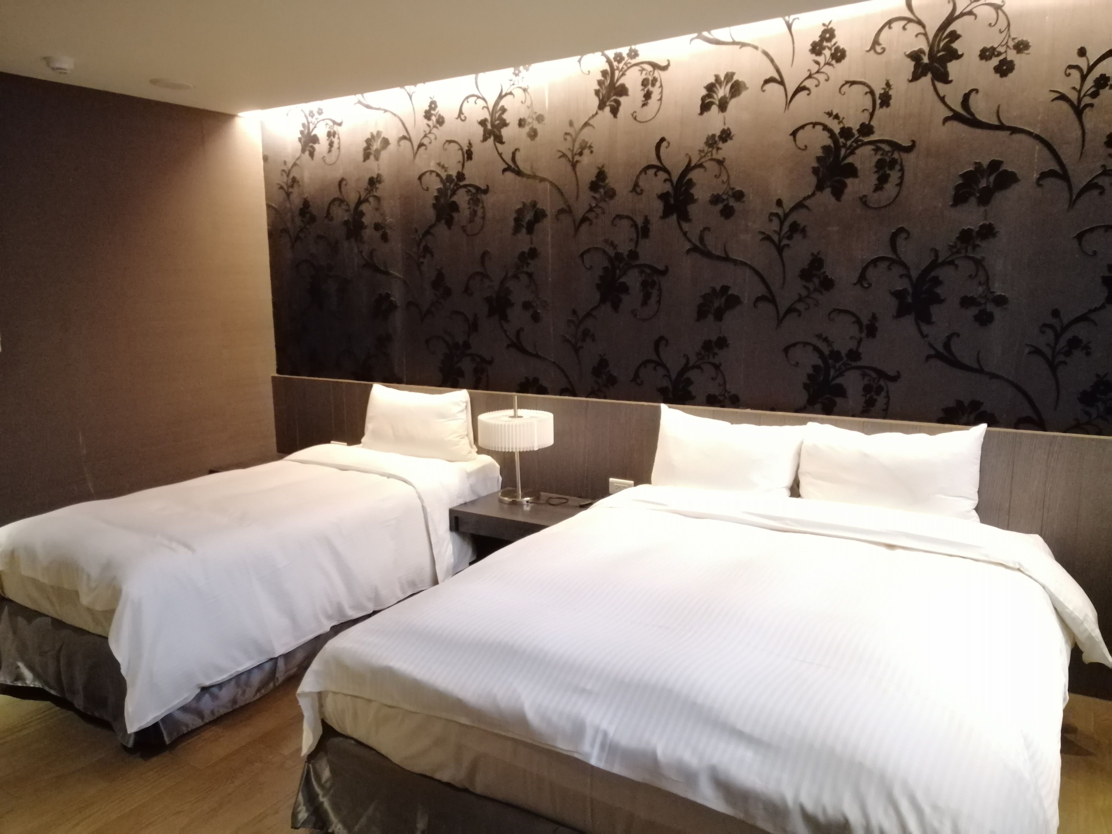 A hotel room with two beds, a nightstand with a lamp, and a floral-patterned headboard.