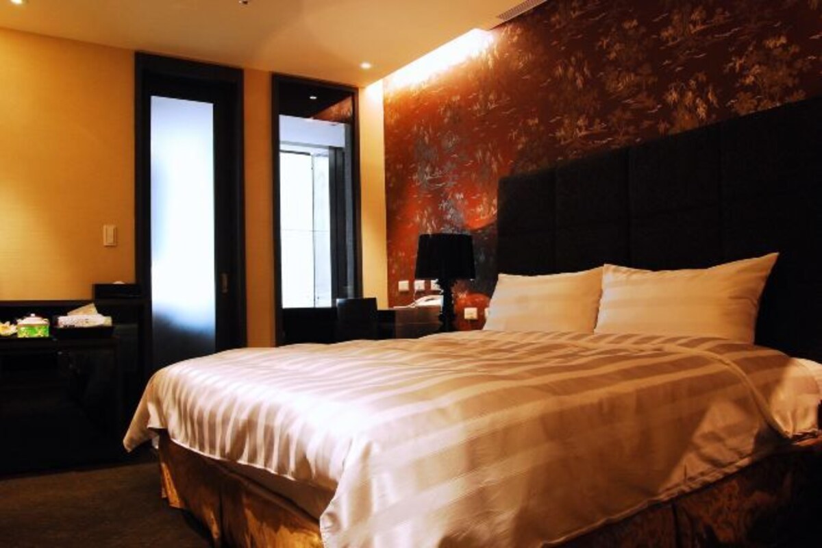 A hotel room with a large bed, a desk with a lamp, and a view of the outside through a door.