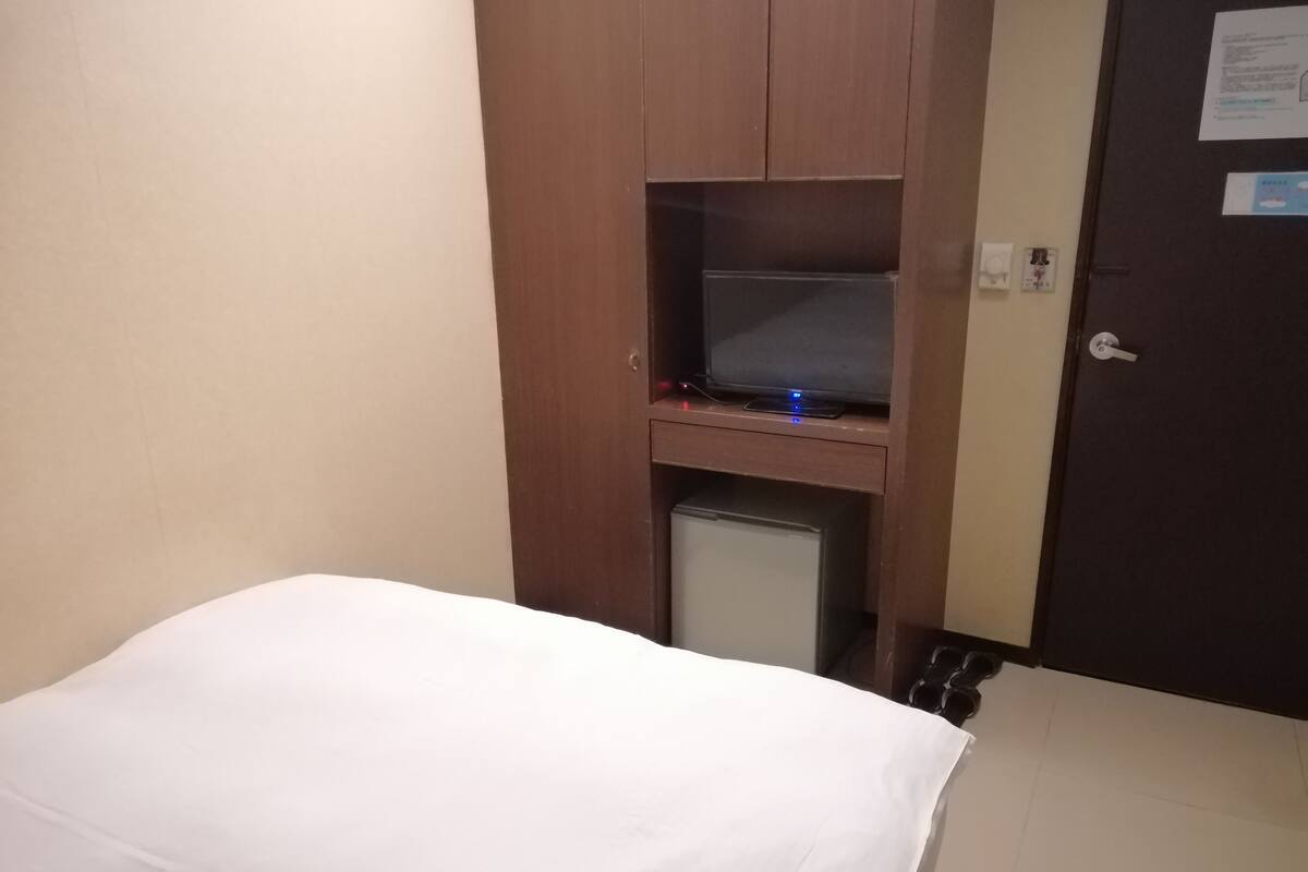 Hotellihuone, jossa on sänky, pieni työpöytä, jossa on televisio, ja jääkaappi.