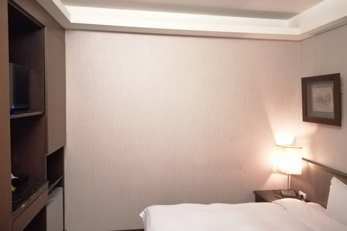 Hotellihuone, jossa on sänky, yöpöytä lampun kanssa, puinen hylly ja seinälle ripustettu taulu.