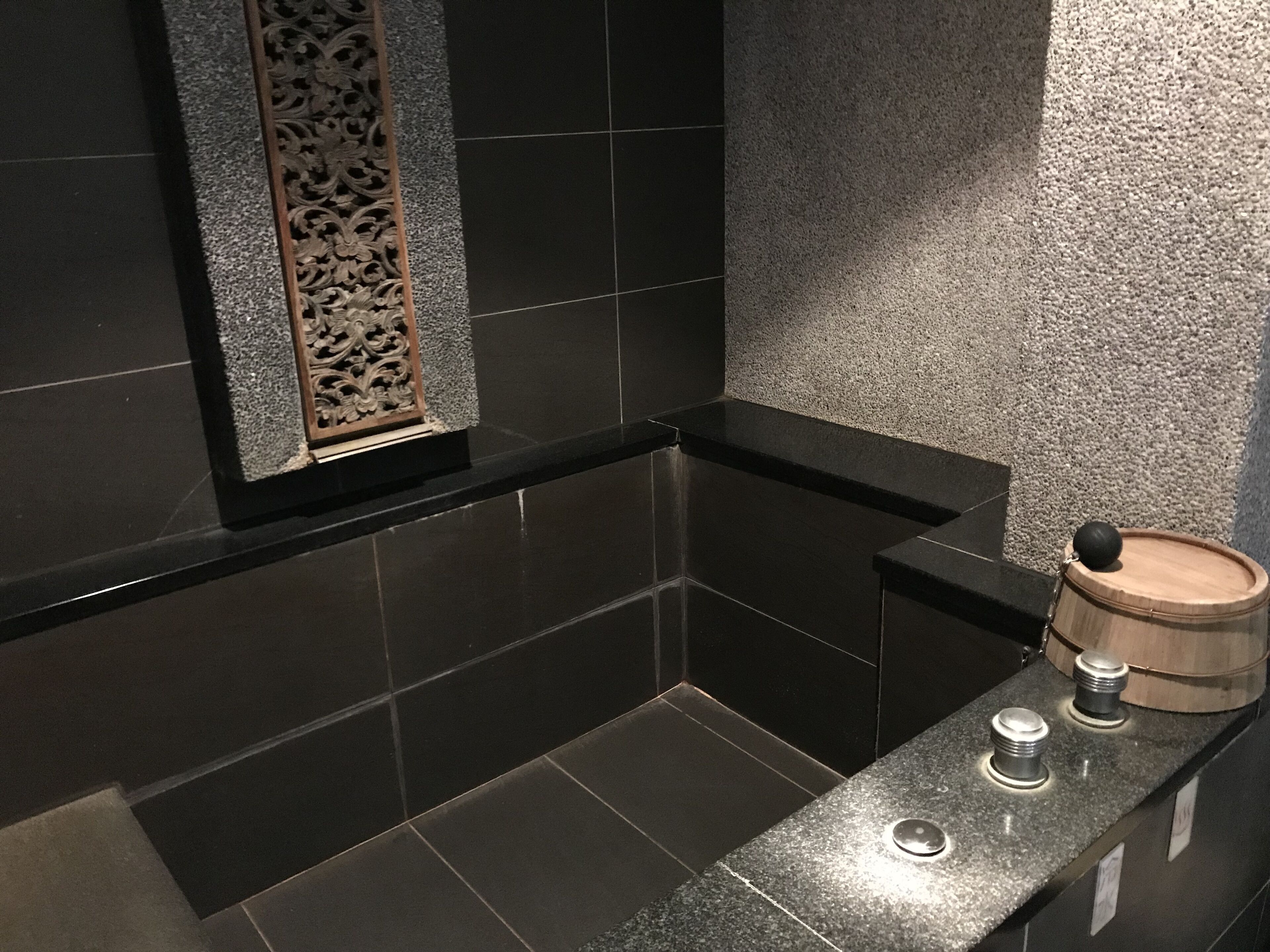 A modern bathroom with dark tiled walls, a wooden decorative panel, and a wooden barrel on the counter.