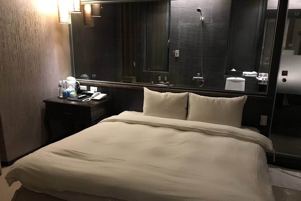 Hotellihuone, jossa on suuri sänky, yöpöytä lampun kanssa, peili ja suihkutila.