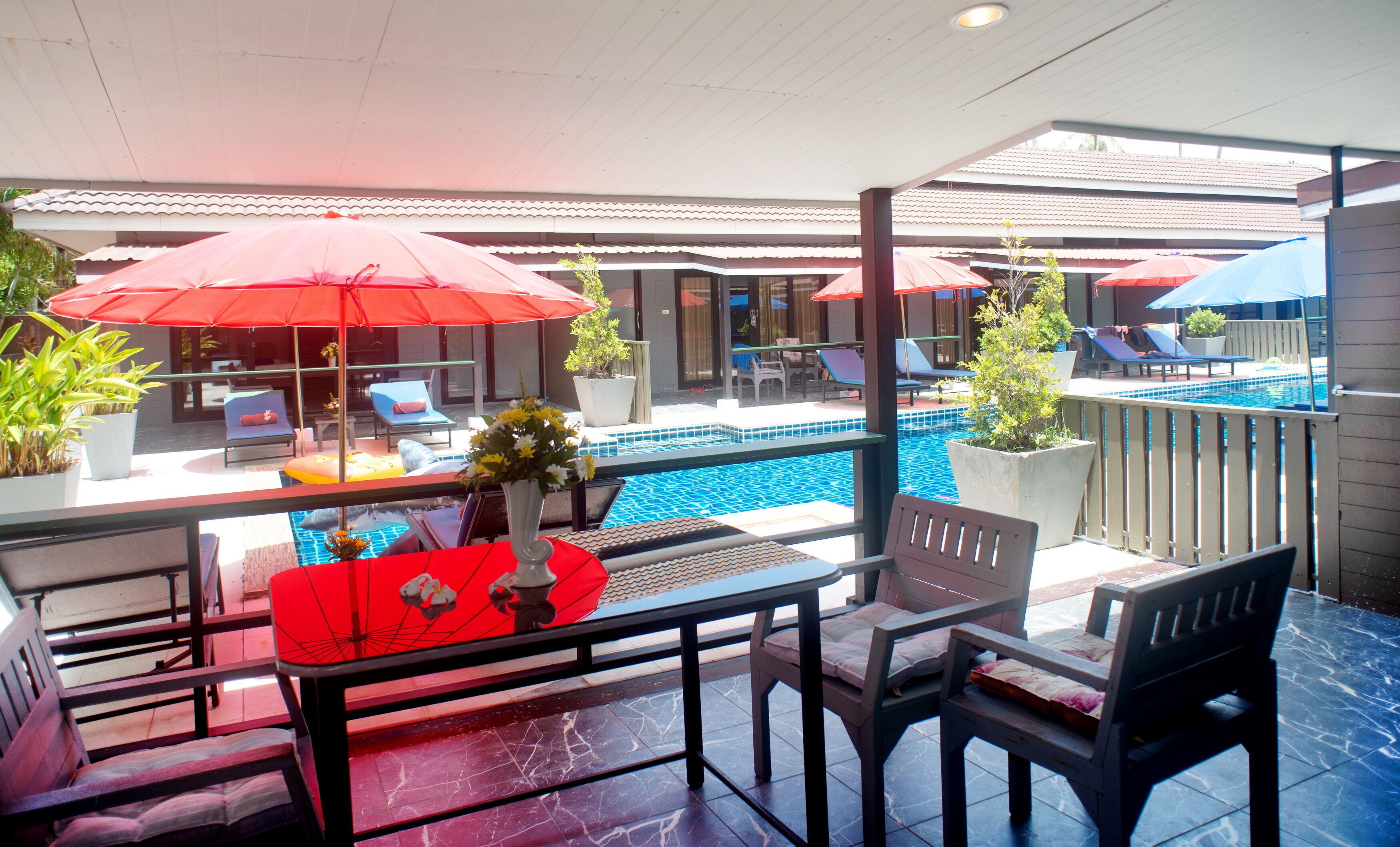 Zona de piscina con sombrillas rojas, sillas de descanso y una mesa de comedor con sillas.