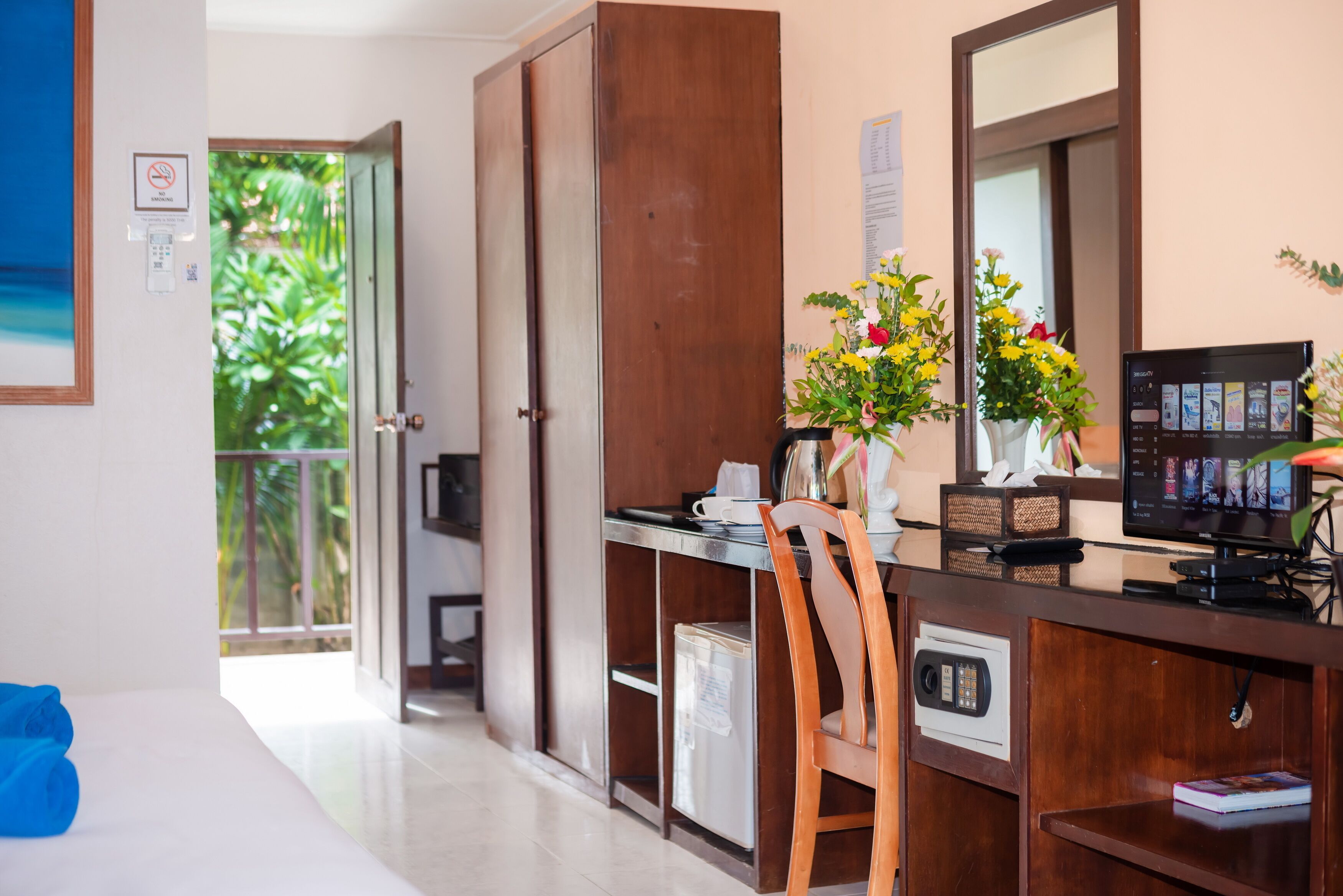 A hotel room with a wooden desk, a chair, a small refrigerator, a microwave, a coffee machine, a TV, and a flower vase.