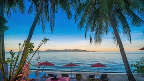 A beachside lounge with palm trees, red umbrellas, and a view of the ocean.