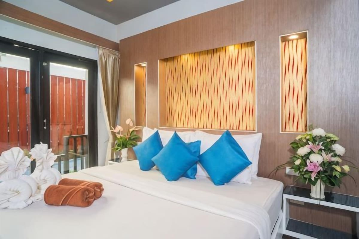 A hotel room with a bed, pillows, a nightstand, a vase of flowers, and a view of a wooden wall and a door.