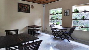 A dining area with wooden tables and chairs, a ceiling fan, a painting on the wall, and a window with potted plants.