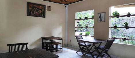 A dining area with wooden tables and chairs, a ceiling fan, a painting on the wall, and a window with potted plants.