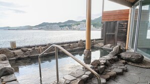 Una piscina de piedra con barandillas de metal, rodeada de rocas y una estructura de madera, con vistas a una masa de agua y edificios a lo lejos.