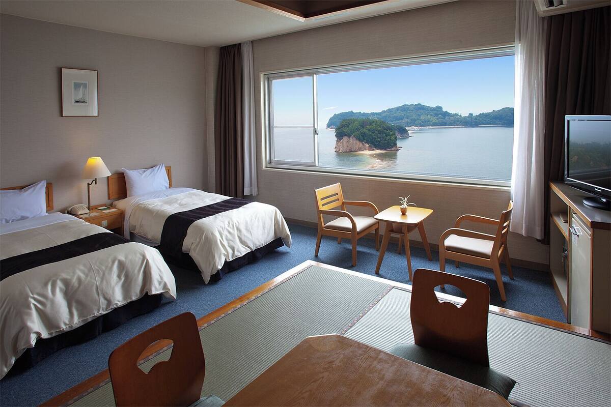 A hotel room with two beds, a TV, a small table, and a view of a lake and island.