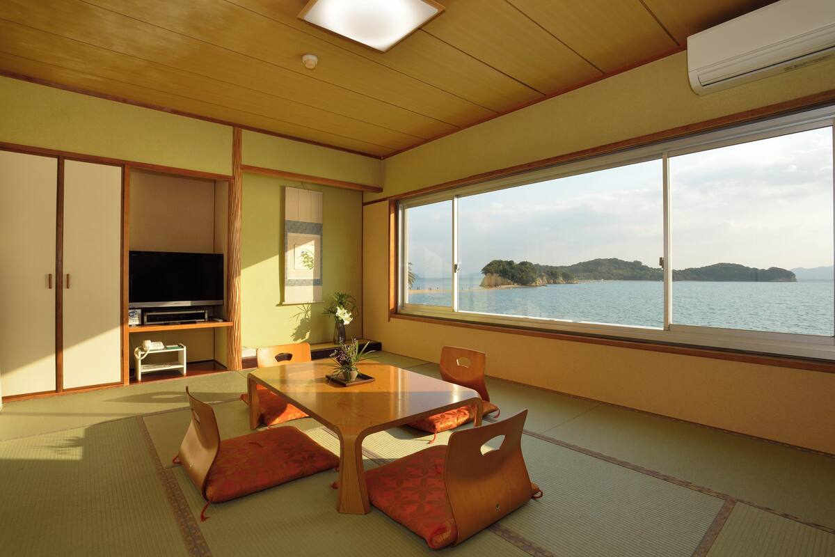 A traditional Japanese room with tatami flooring, a low table, and a large window offering a view of the sea.