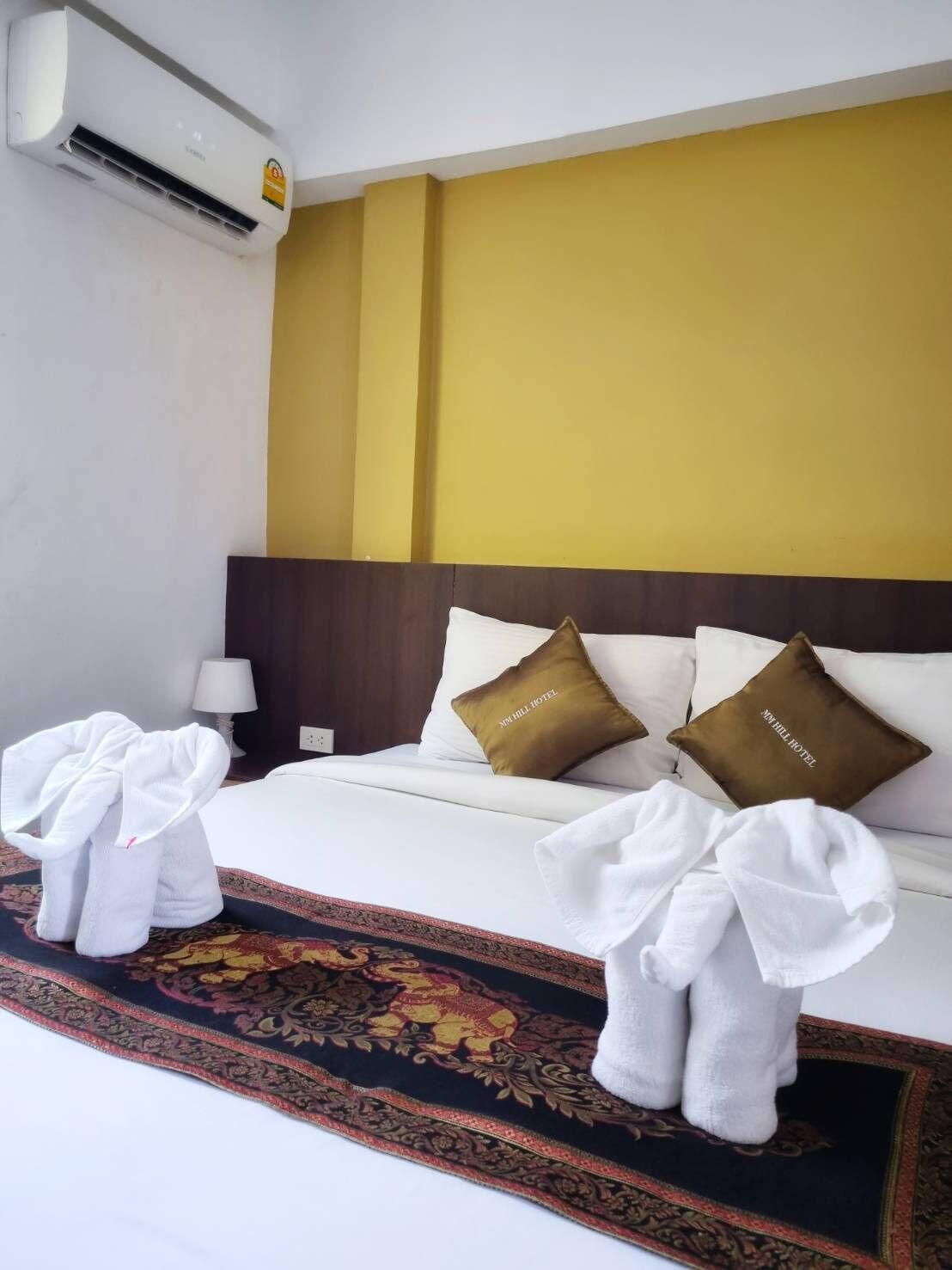 A hotel room with a bed, two pillows with the hotel name, an air conditioner, and two towels on the floor.