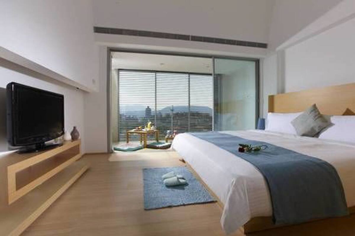 A modern hotel room with a large bed, a television on a wooden shelf, a view of the city through large windows, and a small table with a vase.