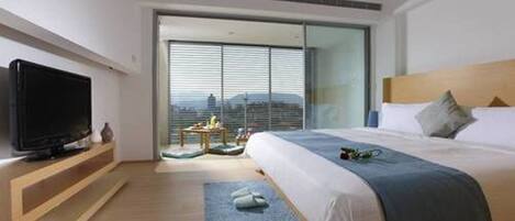 Una habitación de hotel moderna con una cama grande, un televisor sobre un estante de madera, vistas de la ciudad a través de amplias ventanas y una pequeña mesa con un florero.