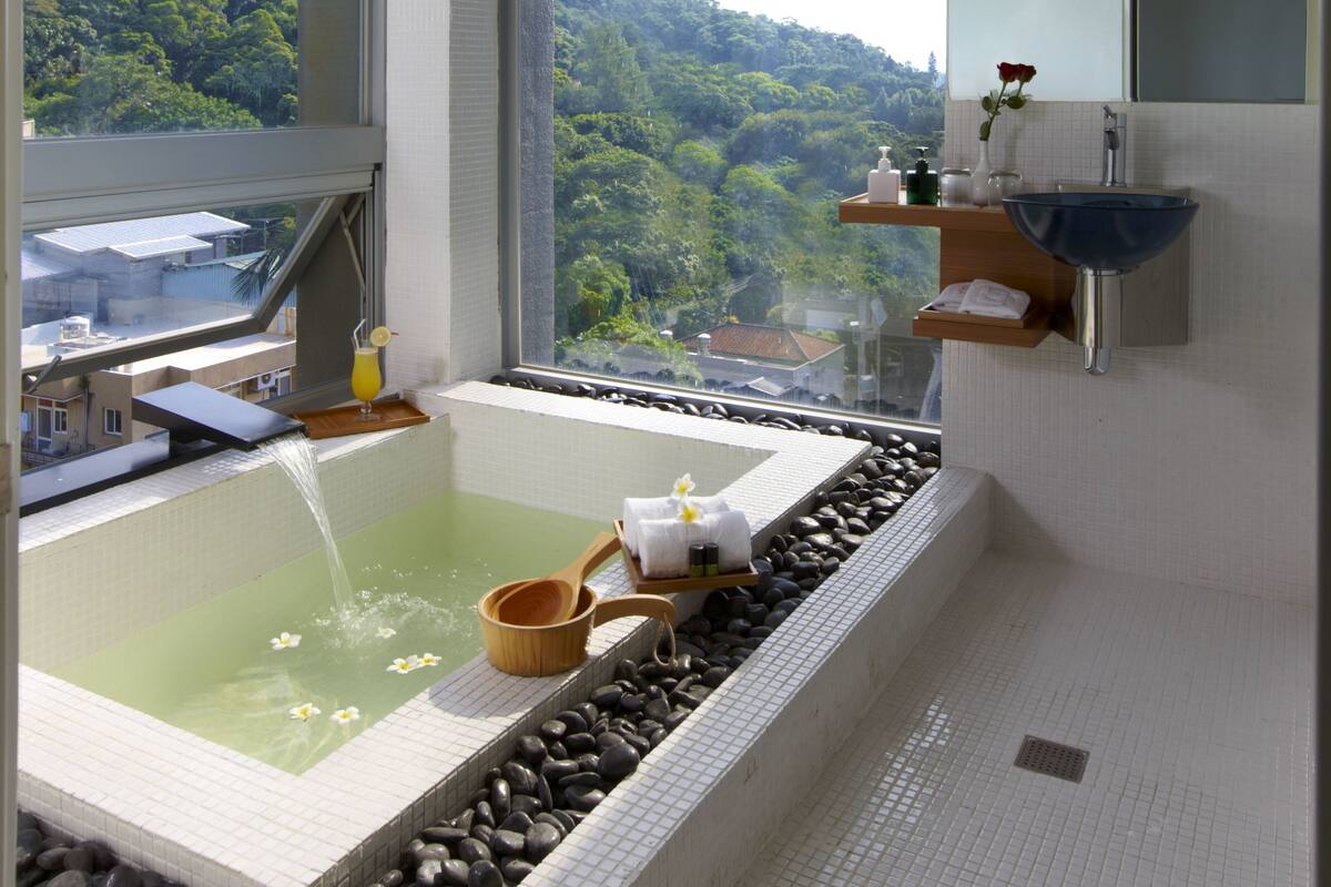 A modern bathroom with a large bathtub, a window with a view of a forest, and a shelf with toiletries.