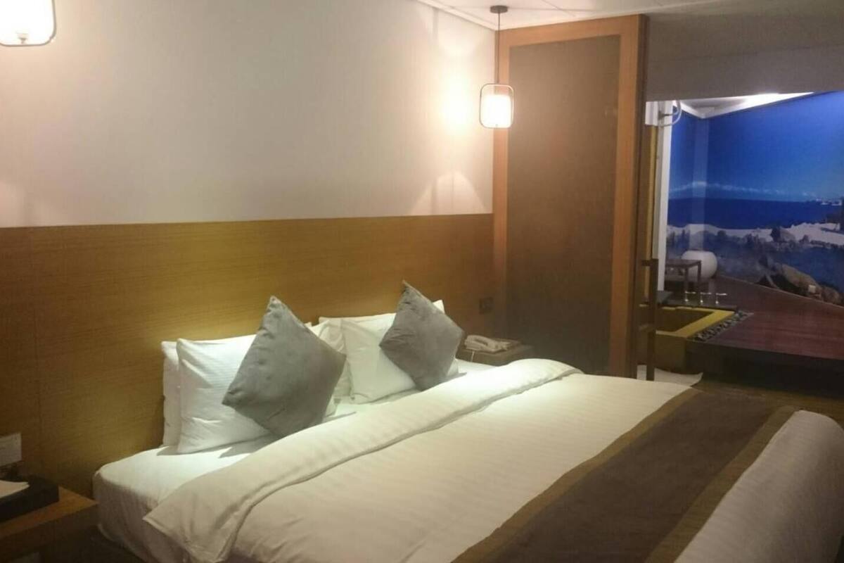 A hotel room with a bed, pillows, a nightstand, a lamp, a desk, and a view of a cityscape through a window.
