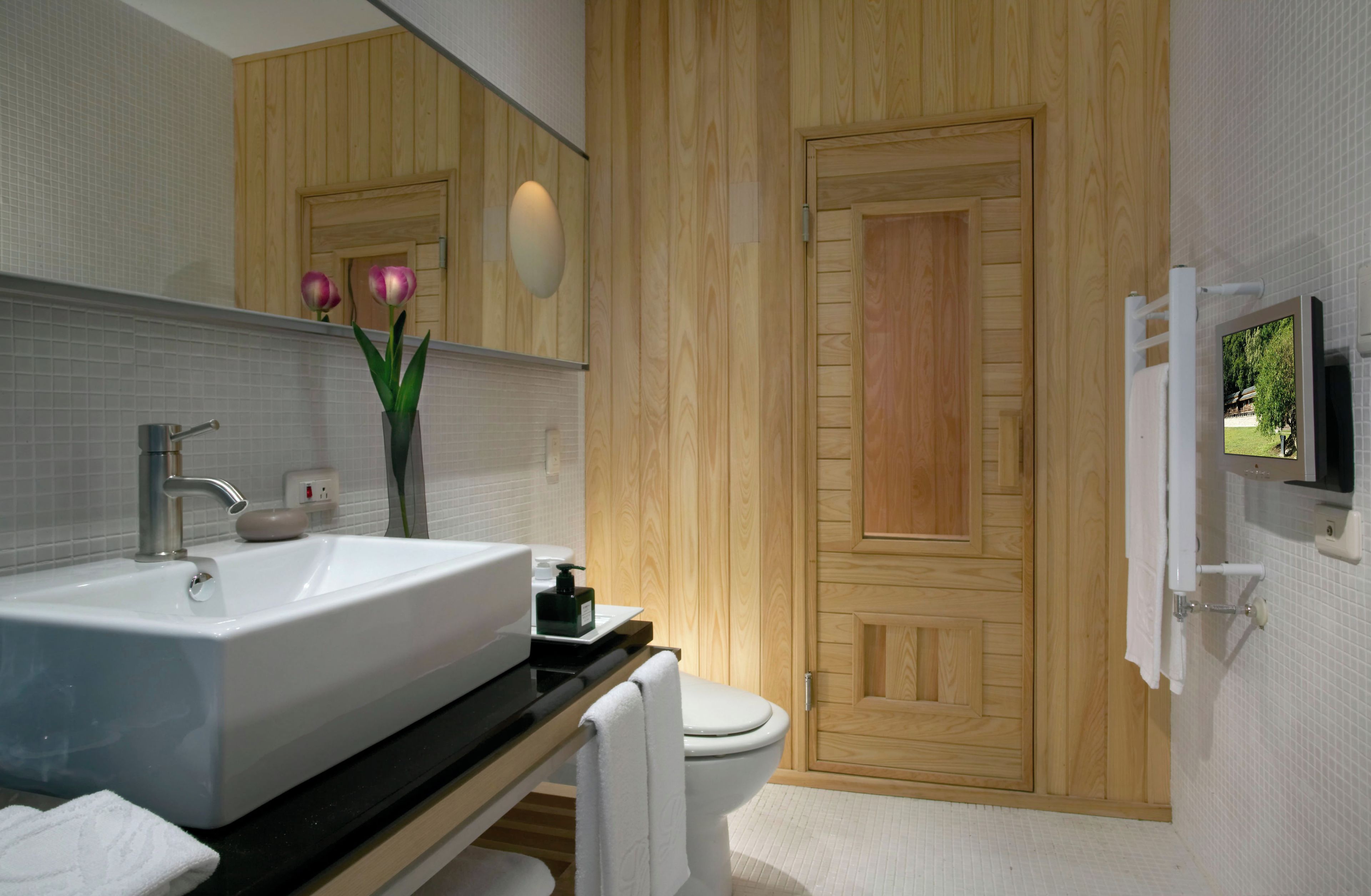 A modern bathroom with a large sink, mirror, wooden door, and a television mounted on the wall.