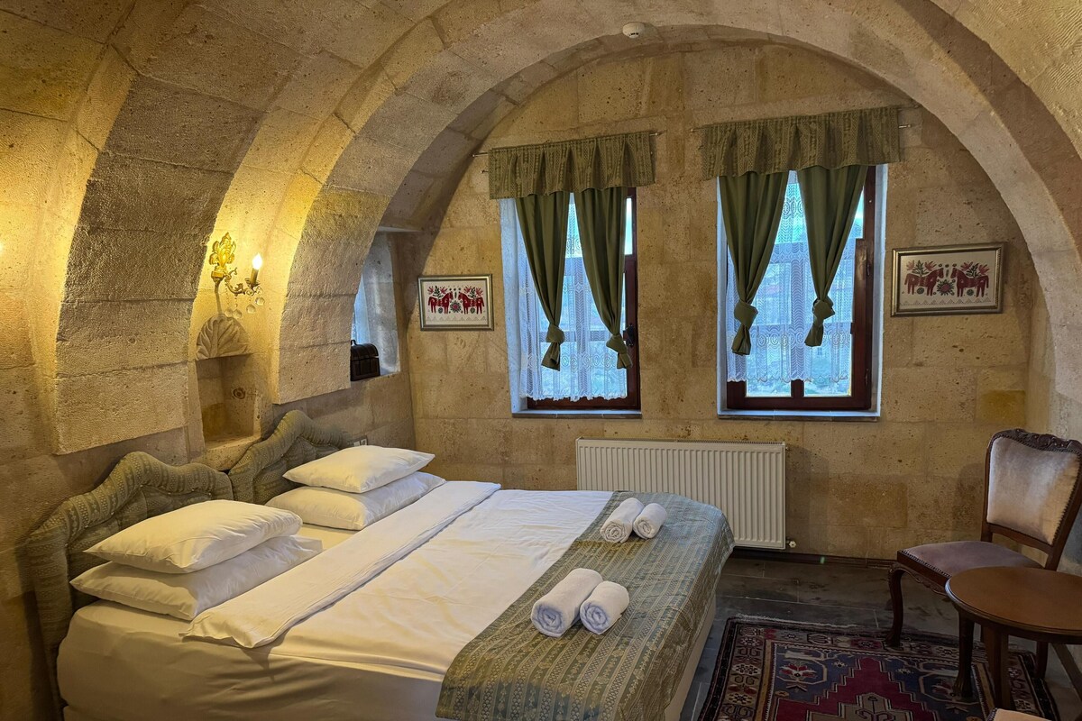 A room with a stone arched ceiling, a bed with white linens, two windows with green curtains, a radiator, and a patterned rug.